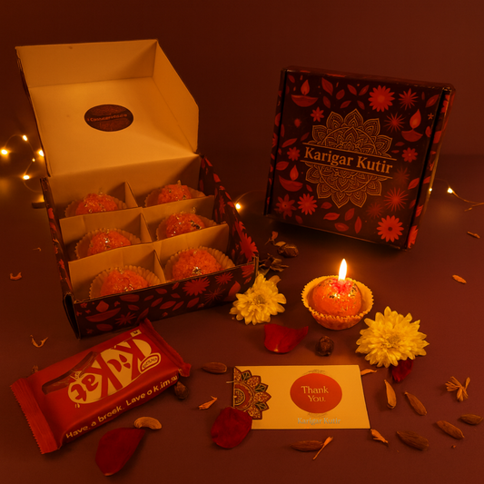 A festive Karigar Kutir gift box display featuring handmade laddoo-shaped candles arranged in an open maroon floral box. One candle is lit beside a thank-you card, surrounded by petals, dry fruits, and flowers. A closed Karigar Kutir box and a KitKat chocolate add a cozy, celebratory touch under warm golden fairy lights.