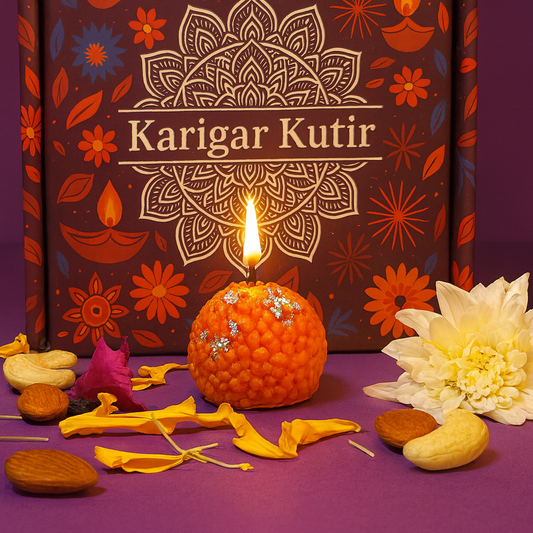 A beautifully lit laddoo-shaped candle with silver varq sits in front of a decorative Karigar Kutir box featuring an intricate mandala design. Surrounded by marigold petals, dry fruits, and a white flower, the scene glows warmly with festive Indian aesthetics and handmade charm.