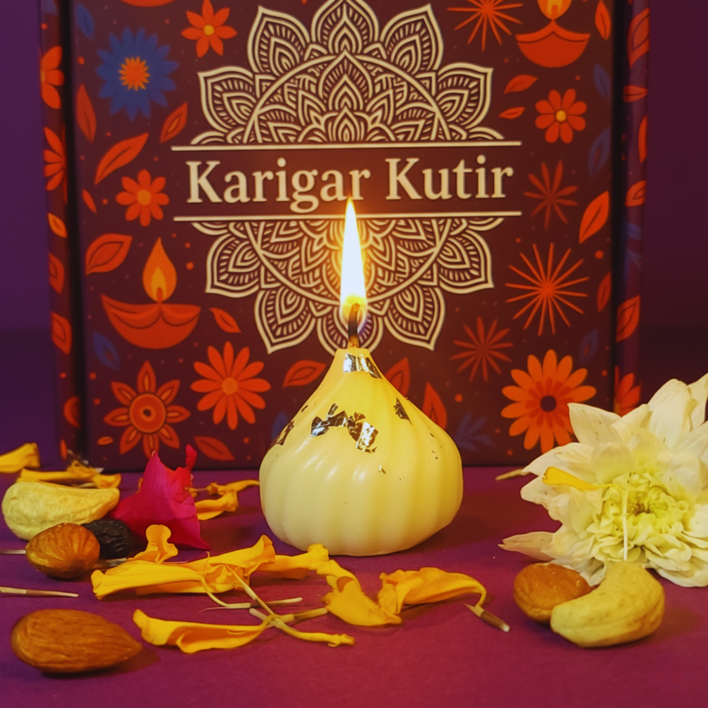 Lit Modak-shaped soy wax candle with silver varq detailing, placed before a Karigar Kutir maroon festive box, surrounded by marigold petals, nuts, and white flowers — symbolizing handmade luxury and Diwali warmth.