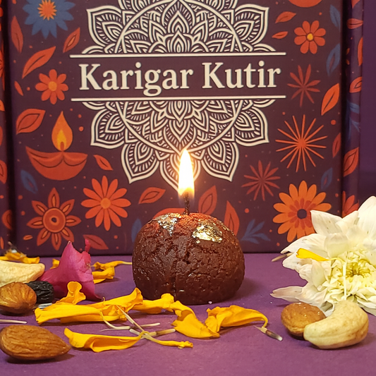 Lit Gulab Jamun-shaped soy wax candle with silver varq detailing, placed before a Karigar Kutir festive box, surrounded by marigold petals, dry fruits, and a white flower — capturing the essence of handmade Diwali luxury.